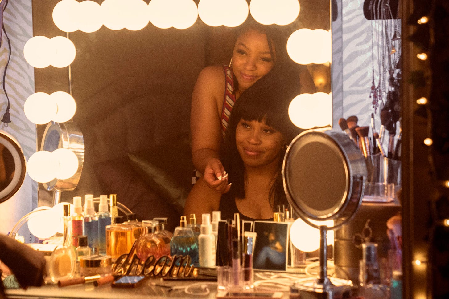 At a bedroom vanity with Hollywood lights around the mirror and cosmetics on the table surface, sits a young Black woman with bangs and shoulder-length hair. Behind her is another young Black woman, whose hair is pulled back, with her arm draped over the shoulder of the woman sitting. They are looking at each other in the mirror with sweet smiles. 