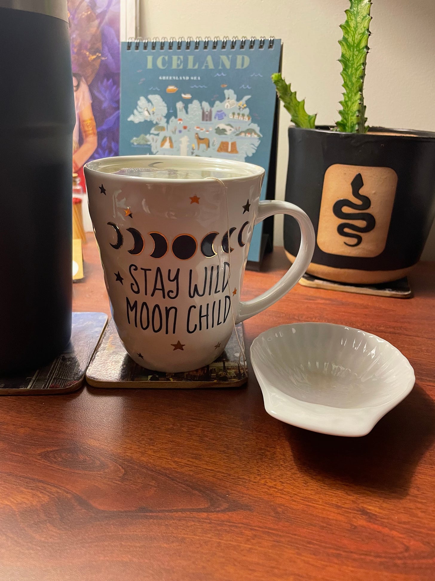 A white ceramic mug with stay wild moon child on it in the foreground, with a calendar and potted plant in the background. 