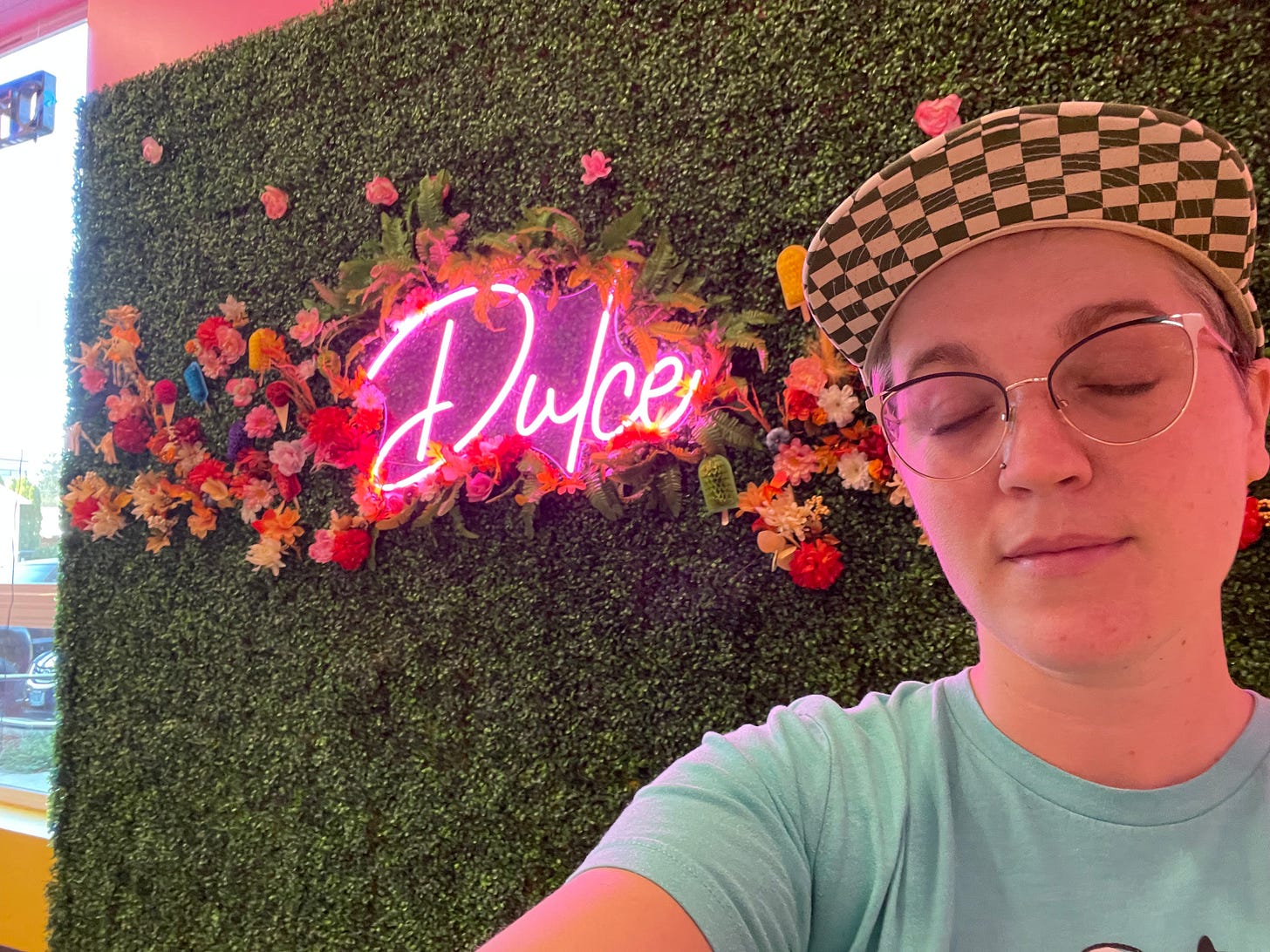 a white person with short hair takes a selfie but has their eyes closed. Behind them is a neon pink sign on a green fake grass background that says dulce