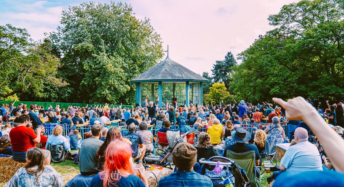 A crowd of people sititng in front of a blue bandstand, with trees around it