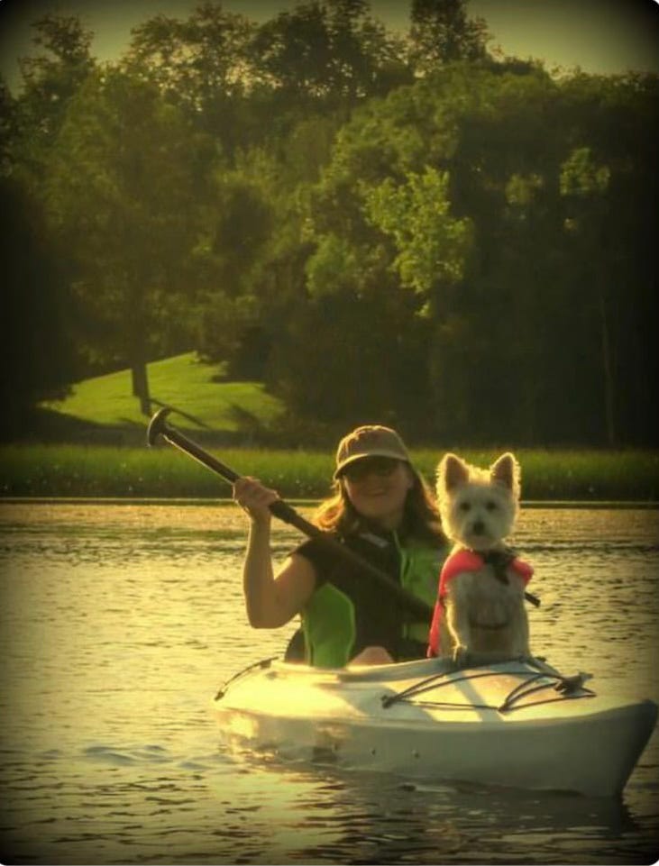 A woman and a small white dog in a kayak on a lake