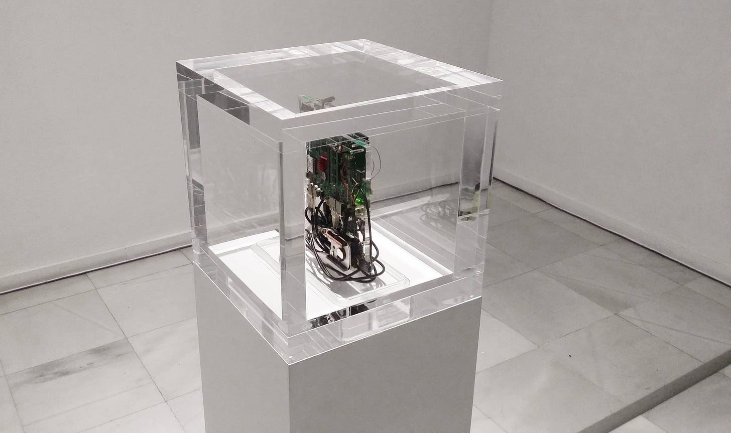 A circuit board in a perspex cube on a plinth in a white walled gallery.