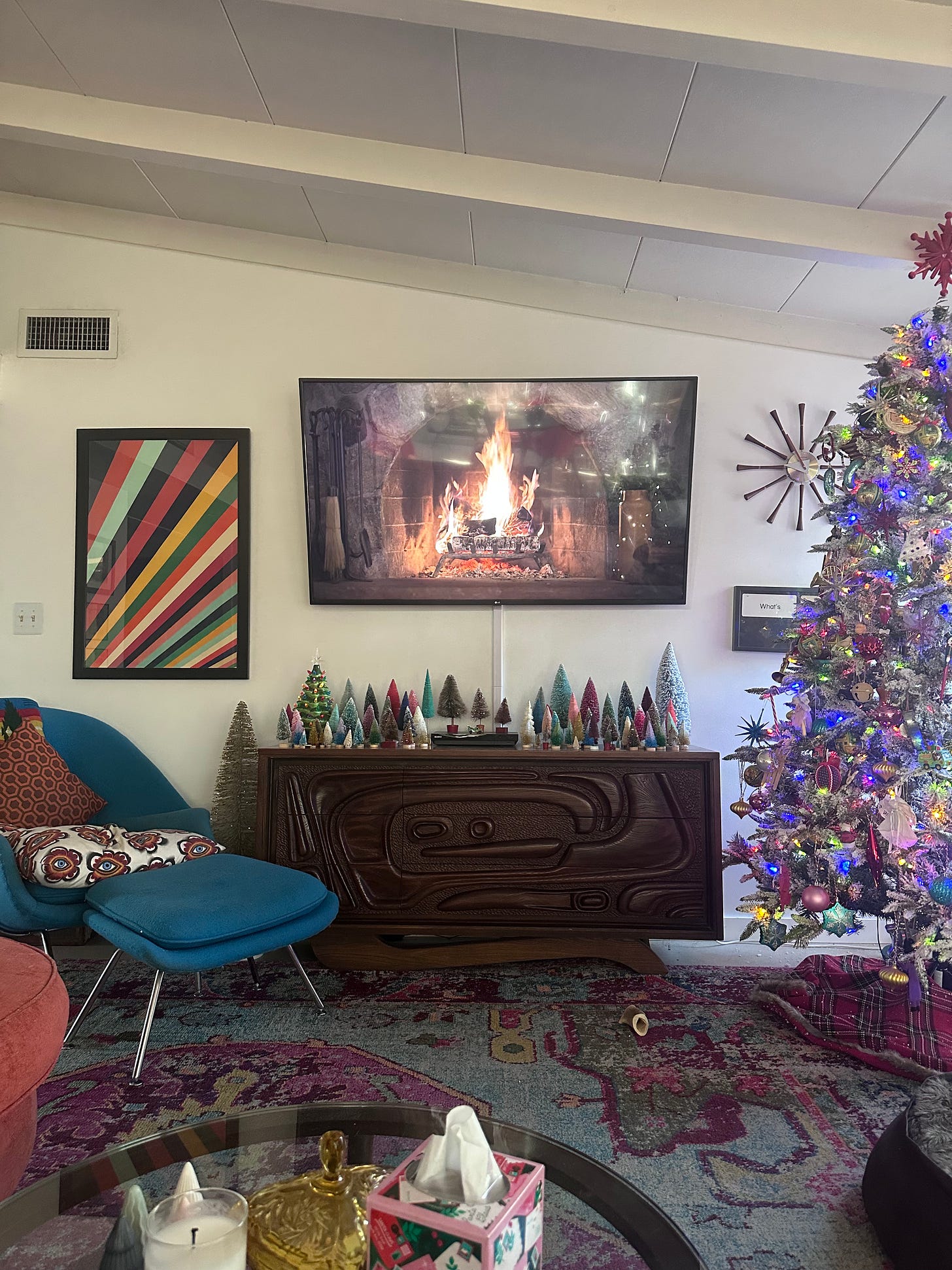 Caissie's living room, decorated for the holidays, featuring her bottle brush tree collection on top of her credenza, beneath a TV screen playing the Yule Log footage