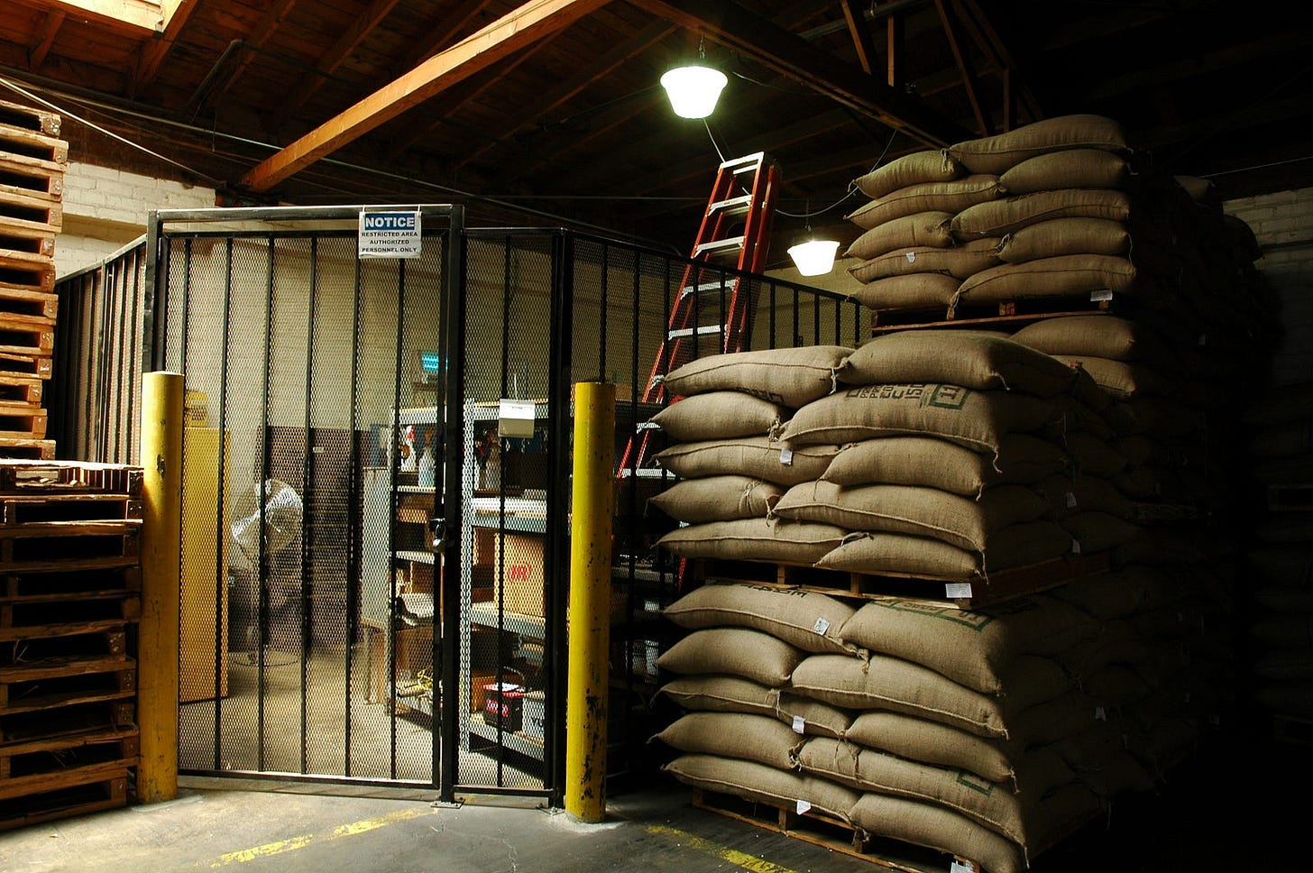 Photo of the darkish inside of a warehouse, showing multiple sacks of something or other stacked up on palettes on the right. There is a stack of empty palettes on the left reaching almost to the ceiling.
