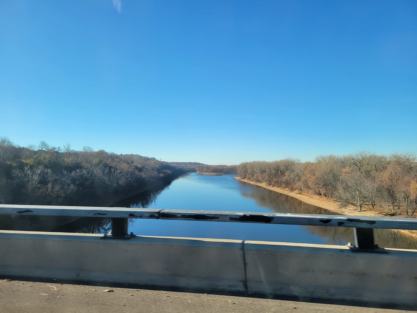 The Mississippi River in St. Paul, MN as seen driving over a bridge. On each sides of the riverbank, there are trees without leaves. The sky is clear and blue. The water is still and reflecting the sky.