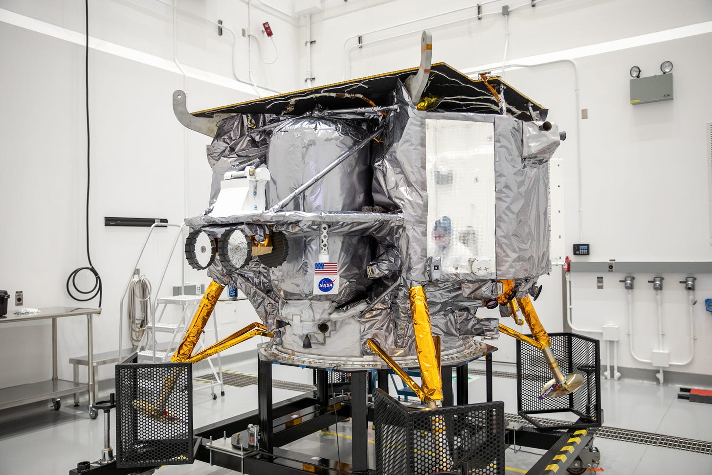 The Peregrine lander sits on a pedestal in a clean room. The masked face of a worker is  reflected in the lander's shiny foil surface.