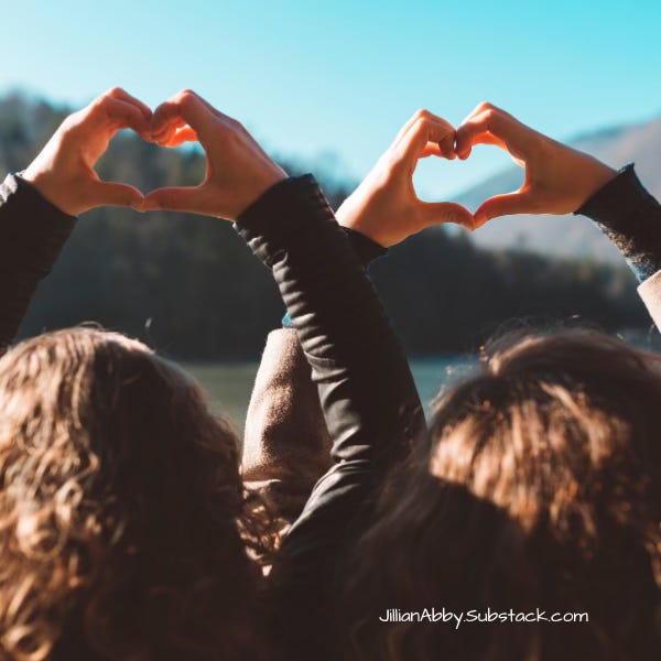 Two women with long brown hair make a heart sign with their hands while looking at a mountain range in the distance.