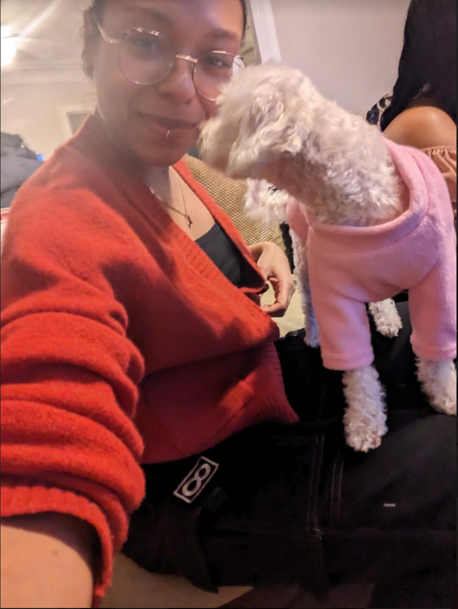 Nathalie takes a selfie with a white dog wearing a pink pullover. Nathalie is wearing a burnt orange cardigan slightly exposing her black sleeveless top