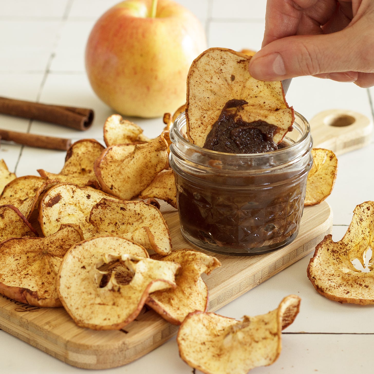 hand dipping apple chip into dark colored dip with more chips and apple and cinnamon stick.