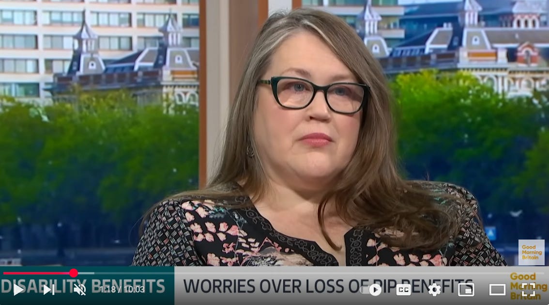 ITV's Good Morning Britain Studio with London Backdrop, Yolanda Barker with long brown/grey hair, green glasses, blue eyes - good skin - if I do say so myself! Black dress with white and peachy coloured flowers and geometric shapes.  Sat in powerchair being interviewed with regards to potential cut in PIP