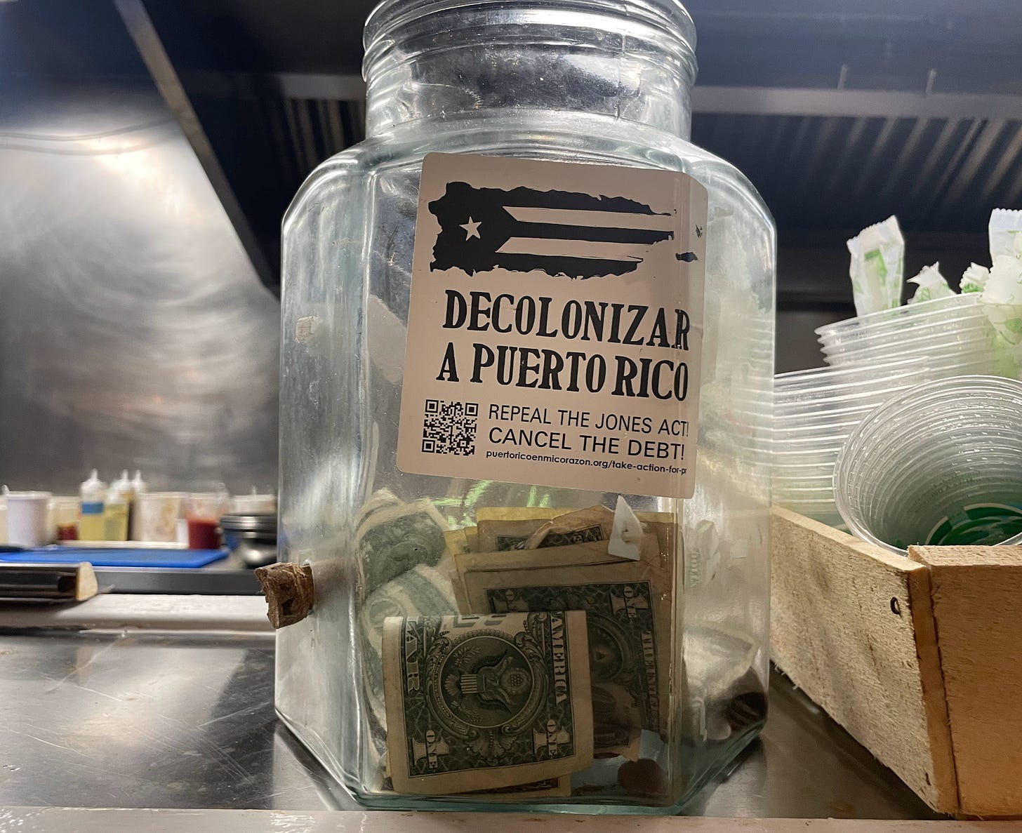 A tip jar in a restaurant is covered with a sticker that reads "Decolonizar a Puerto Rico", with a QR code and link