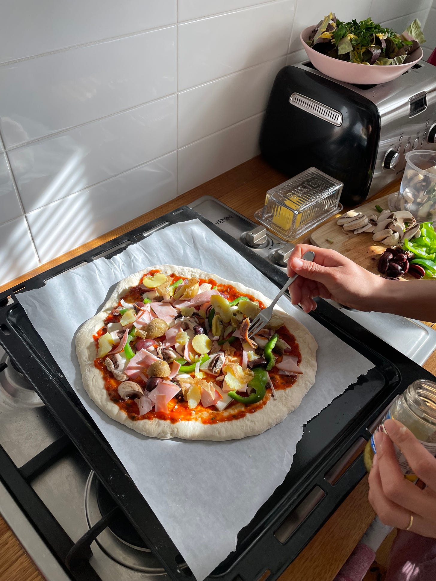Uncooked pizza topped with a tomato sauce, vegetables on a baking tray, with someone putting olives on top.