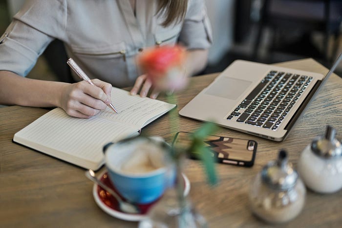 A woman writing something in a notebook while the laptop, also on the table, remains unused for the time being