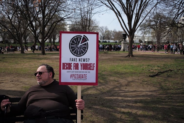 A man sits on a bench in a park holding a sign that reads: fake news decide for yourself Pizzagate. A man sits on a bench in a park holding a sign that reads: fake news decide for yourself Pizzagate.