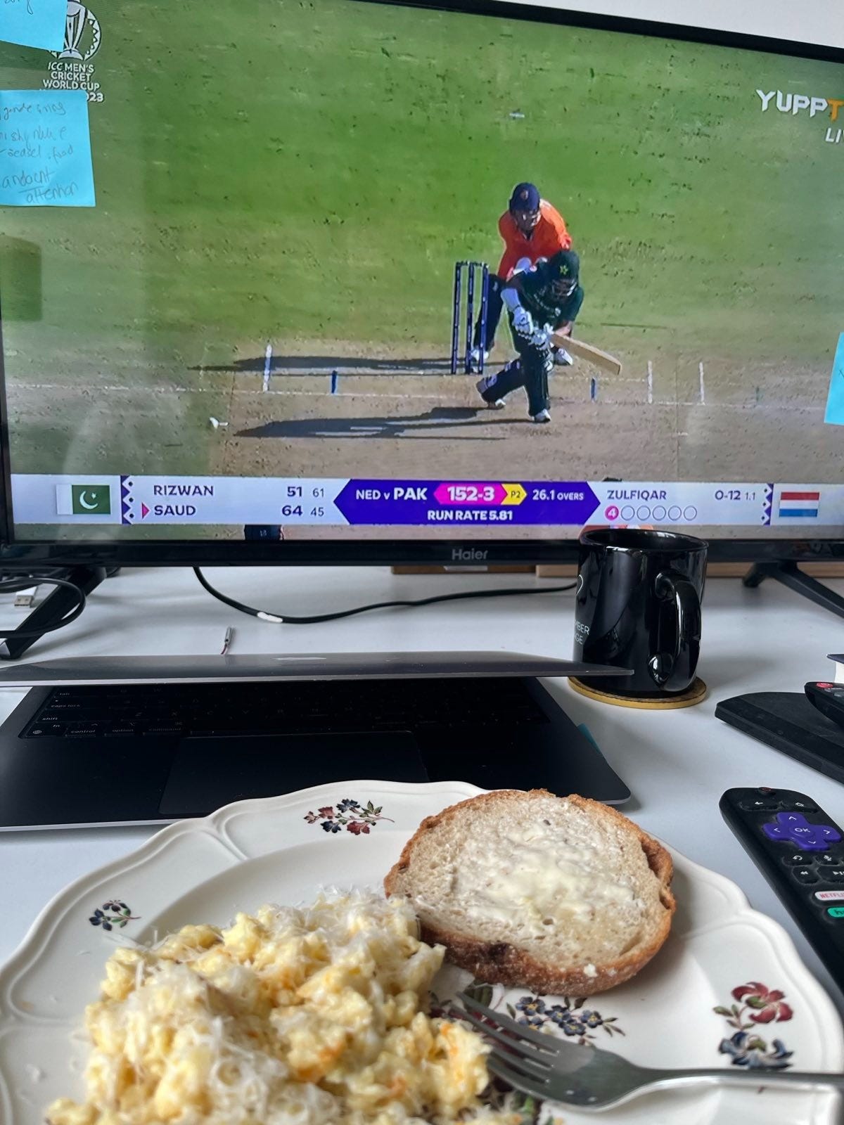 A television screen shows a cricket match. in front of it is a plate of scrambled eggs and toast.