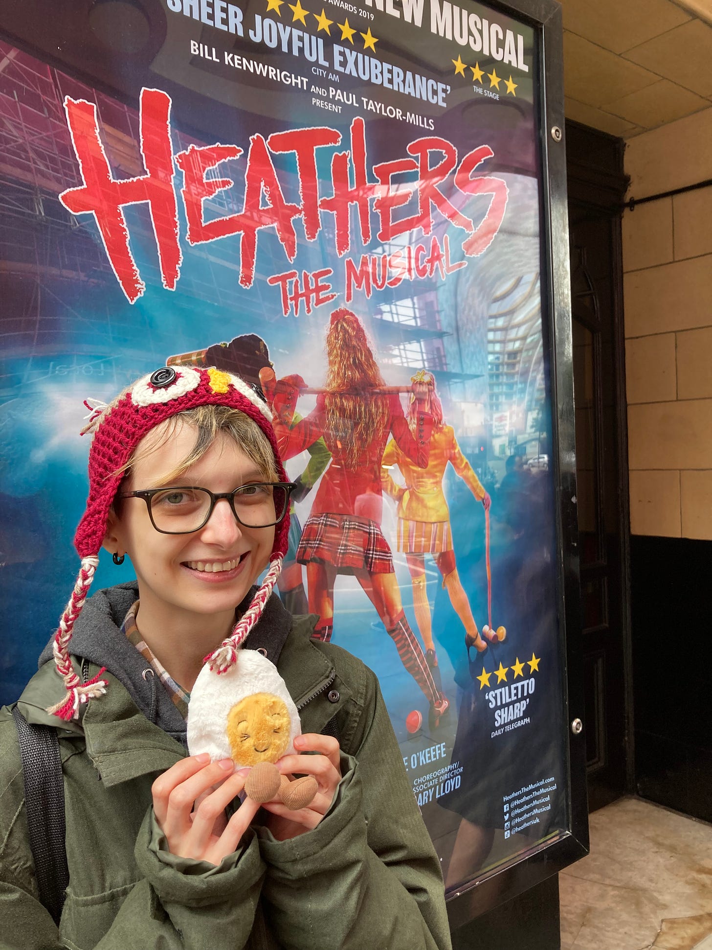 Photograph of Beck holding a soft toy boiled egg outside, in front of a poster for Heathers the Musical. Beck is smiling and they are slightly windswept and goofy looking.