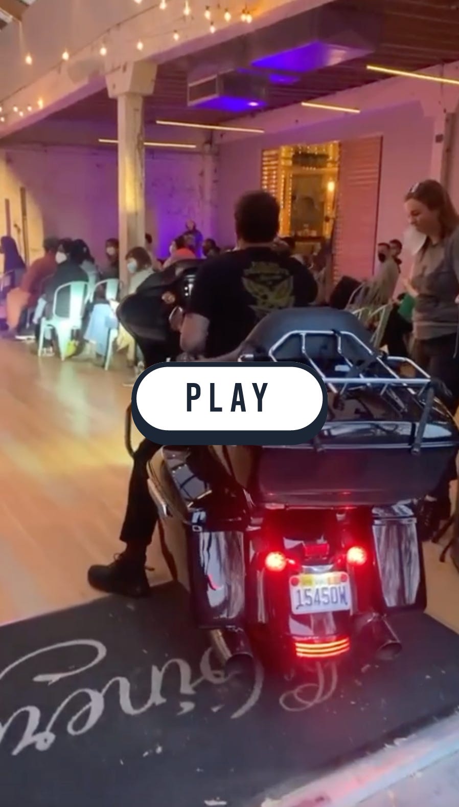 A screenshot of a video with a button that says "Play" on it. Screenshot is of Derrick is riding the motorcycle into a venue.