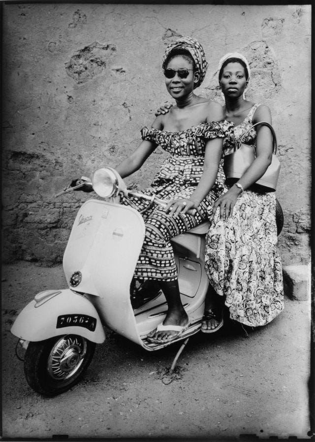 Two women from Mali pose on a white Vespa scooter in a black and white photo