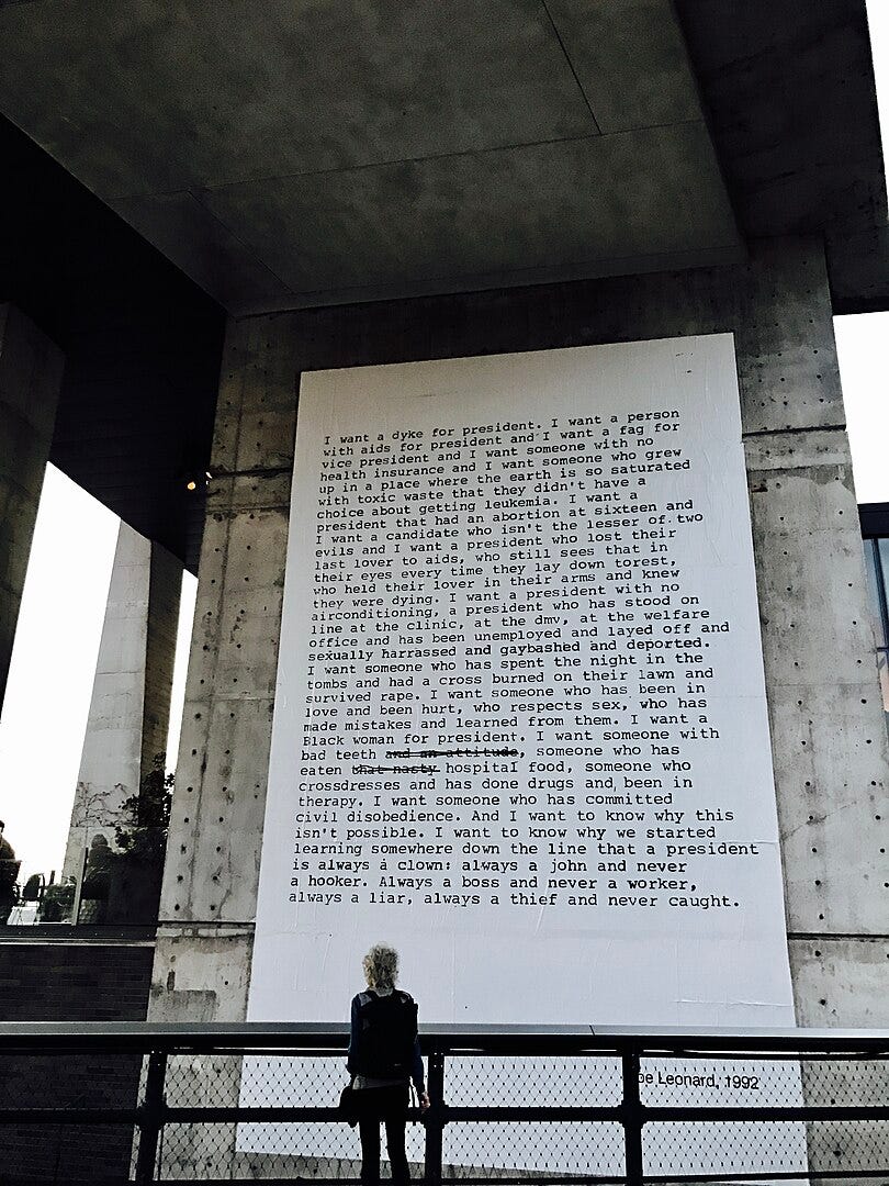 A person stands in front of a huge white banner with black typed text. The banner is installed on a huge wall under a bridge.