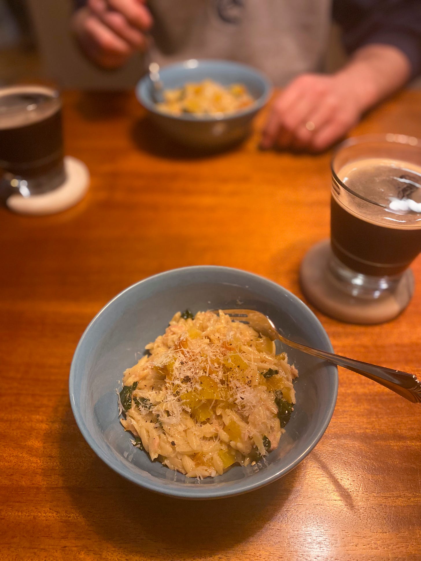 Two blue bowls of the orzo dish described above, dusted with parmesan and black pepper. Above and to the right of the bowl on coasters are glasses of dark beer. In the background, Jeff's hand rests on the table next to his bowl.