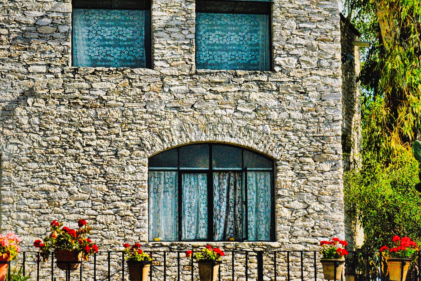 A stone house with two small windows with lace curtains on the second floor and a large arched window on the first floor with a fence and pots of red geraniums in front