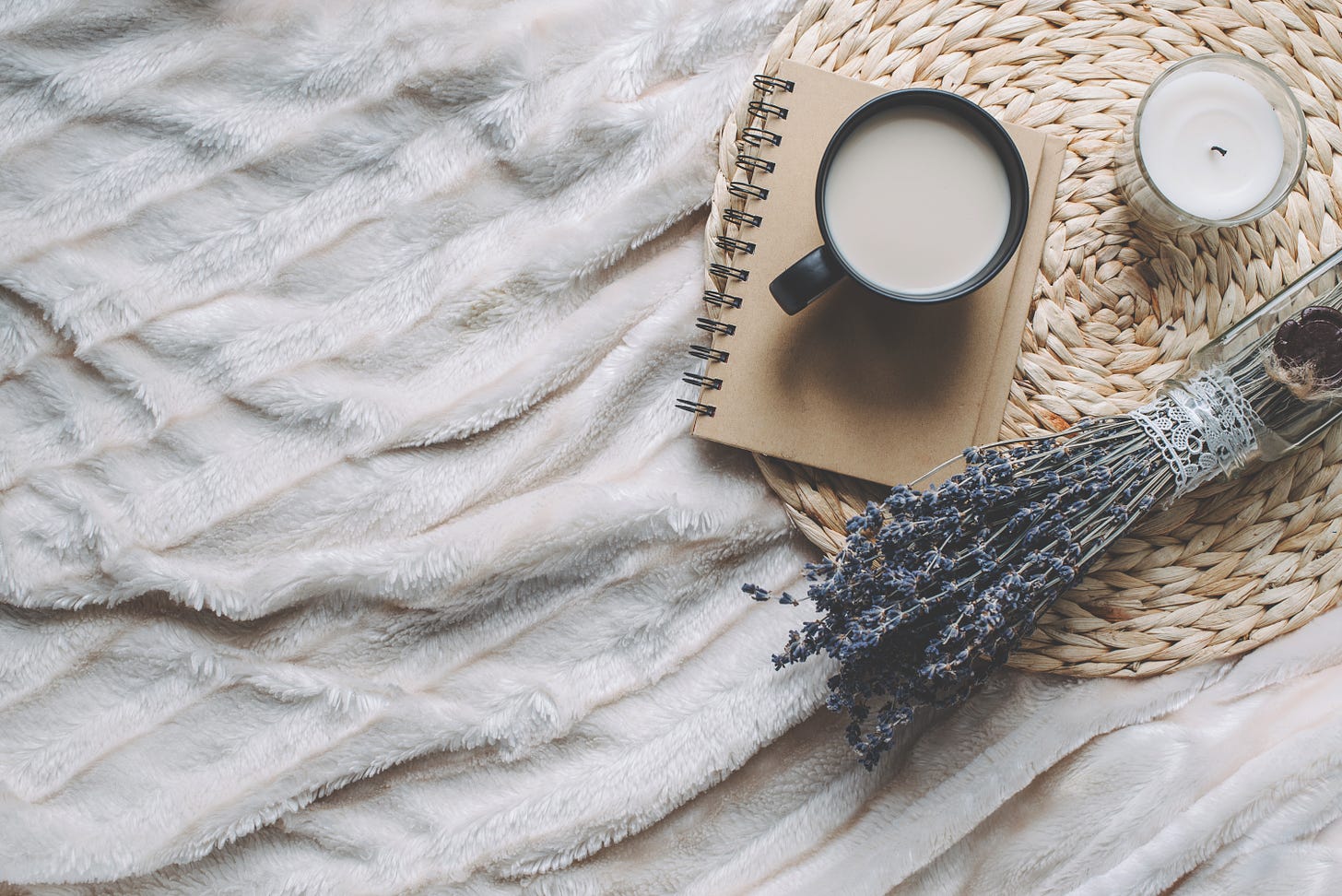 Mug with coffee, notebook, a lavender bunch and a white candle on a wicker tray, over a fluffy blanket.