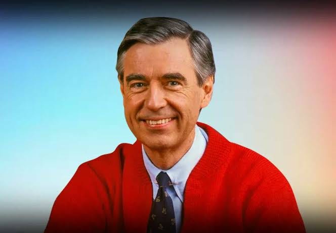 Photo of Fred Rogers wearing a read cardigan sweater blue shirt and navy tie.
