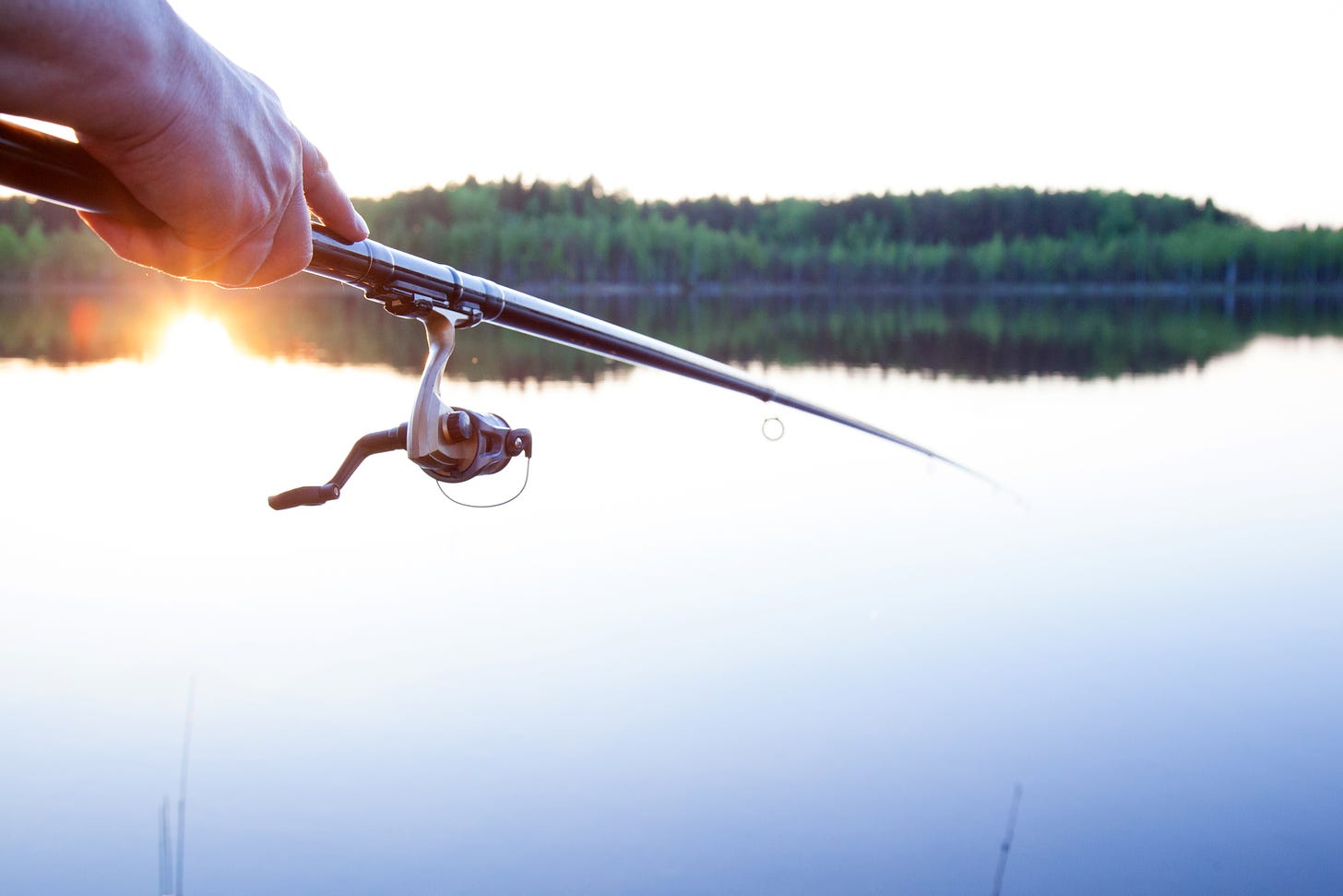 Person's hand holding a fishing pole in front of a large, still lake with a tree line and the sun reflecting on the water