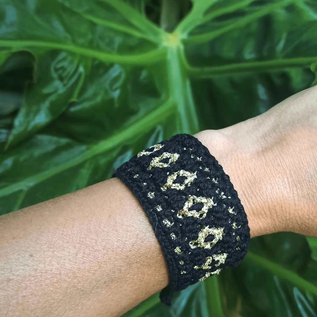 Mosaic crochet bangle-style bracelet in black and golden thread, with geometric pattern, on wrist with big plant leaves in the background.