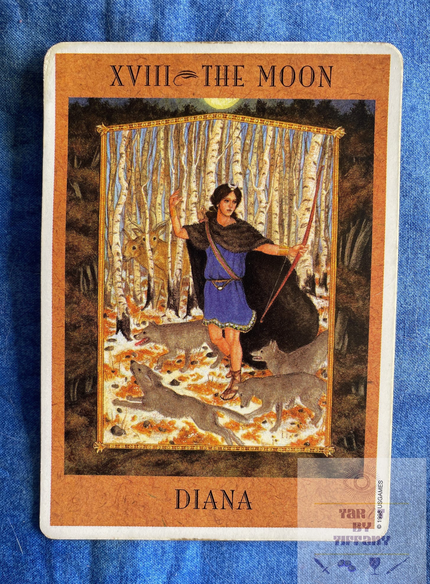 Image of the Moon card from the Goddess Tarot. The card shows the goddess Diana dressed in a blue tunic and a fur cape. She is holding her bow in her left hand and her right hand is raised as if she has just released an arrow. Four gray wolves surround her feet in a circle; they are her wolves. There is snow on the ground and the trees are bare. Behind her are two deer, hiding behind one of the bare tree trunks. The card is laying on a cobalt blue cloth background.