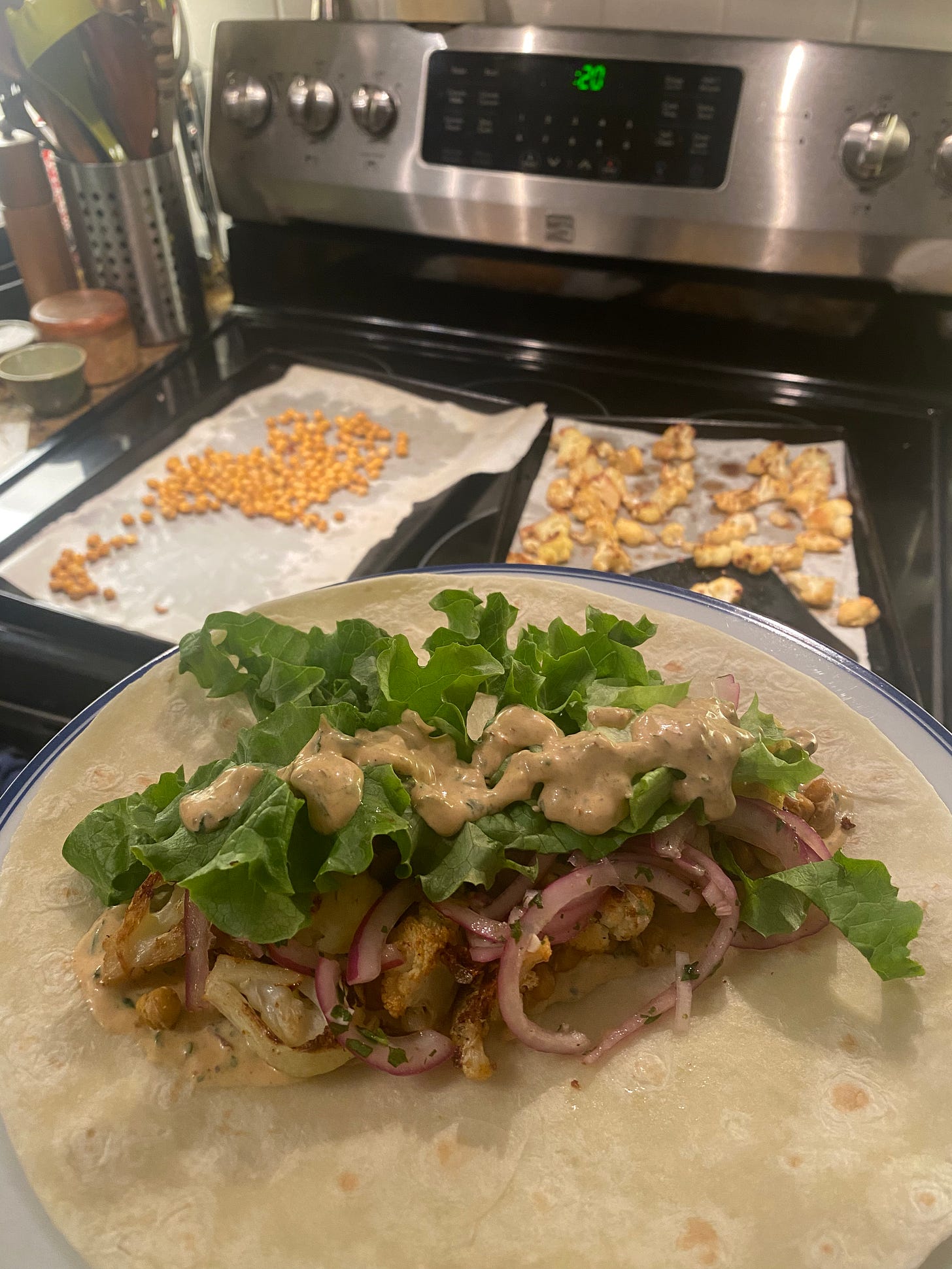 In the foreground on a plate, an open tortilla with the chickpeas, cauliflower, and onions underneath green lettuce and the sauce described above. In the background on top of the stove, the two baking pans of chickpeas and cauliflower are visible.