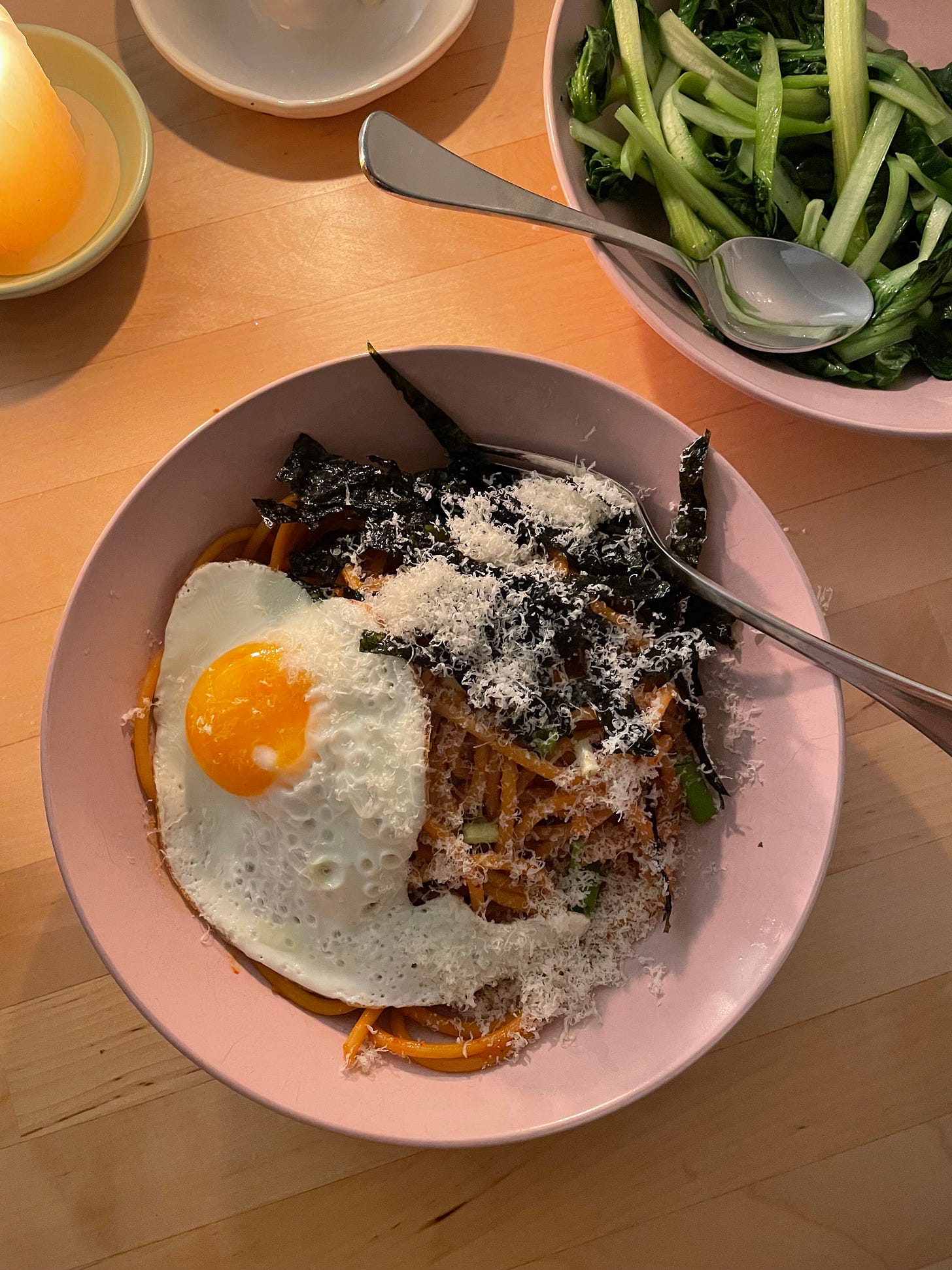 Bowl of kimchi linguini topped with roasted seaweed, parmesan and a fried egg. Garlicky greens in a bowl nearby.