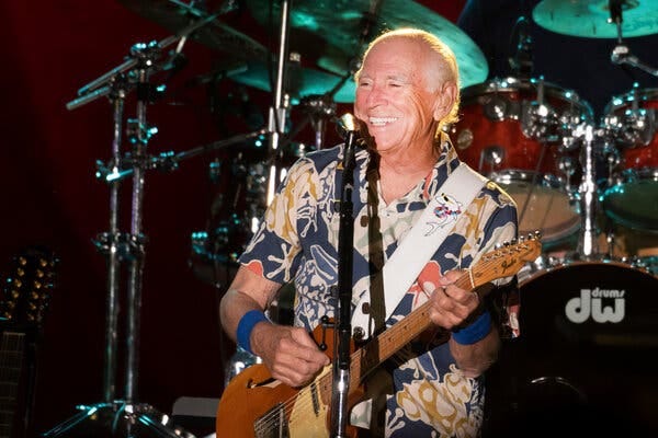 An older man with thinning gray hair wears a Hawaiian shirt as he strums a Fender telecaster electric guitar on a concert stage.