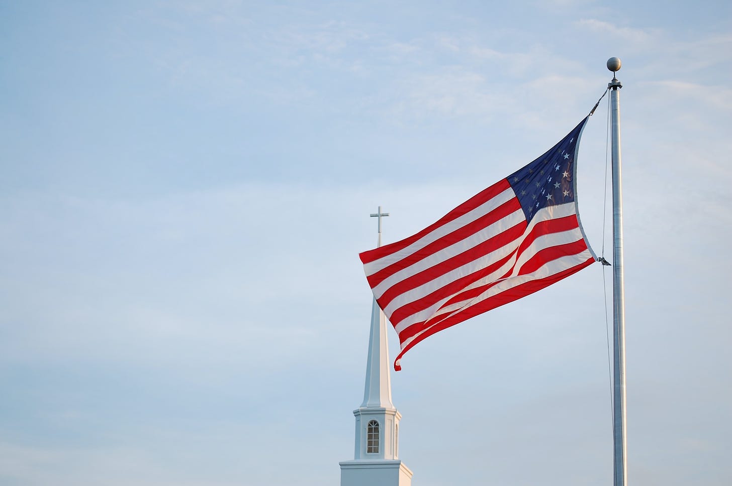 Large American flag flying in front of a church steeple with a cross on top.
