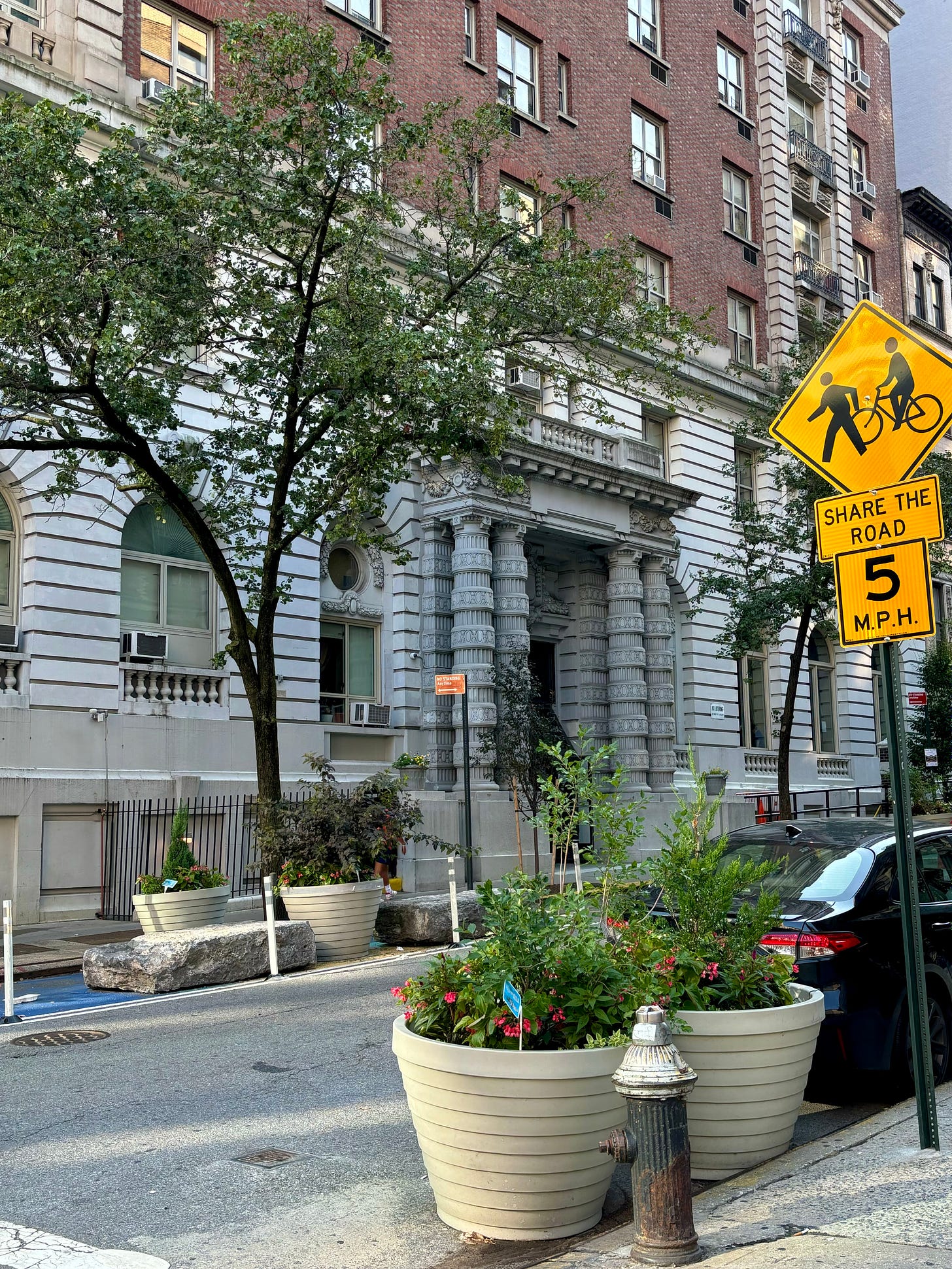 A large red brick apartment building. The facade over the first two floors are stone and the entrance is flanked by ornately carved columns. To the right is a sign warning of of pedestrians and bikers as the street appears closed to traffic to allow for the residents to enjoy it.