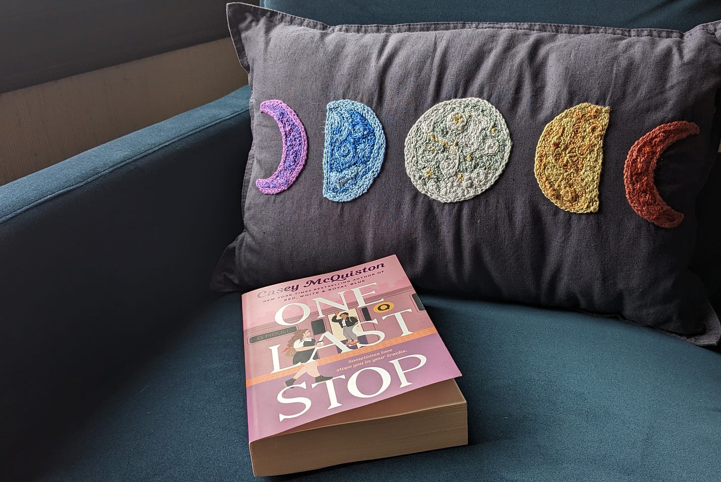 On a midnight blue velvet armchair is a dark grey pillow beautifully embroidered with five phases of the moon, in front of which is a pastel pink-and-purple paperback book with all-caps white letters that read 'ONE LAST STOP'.
