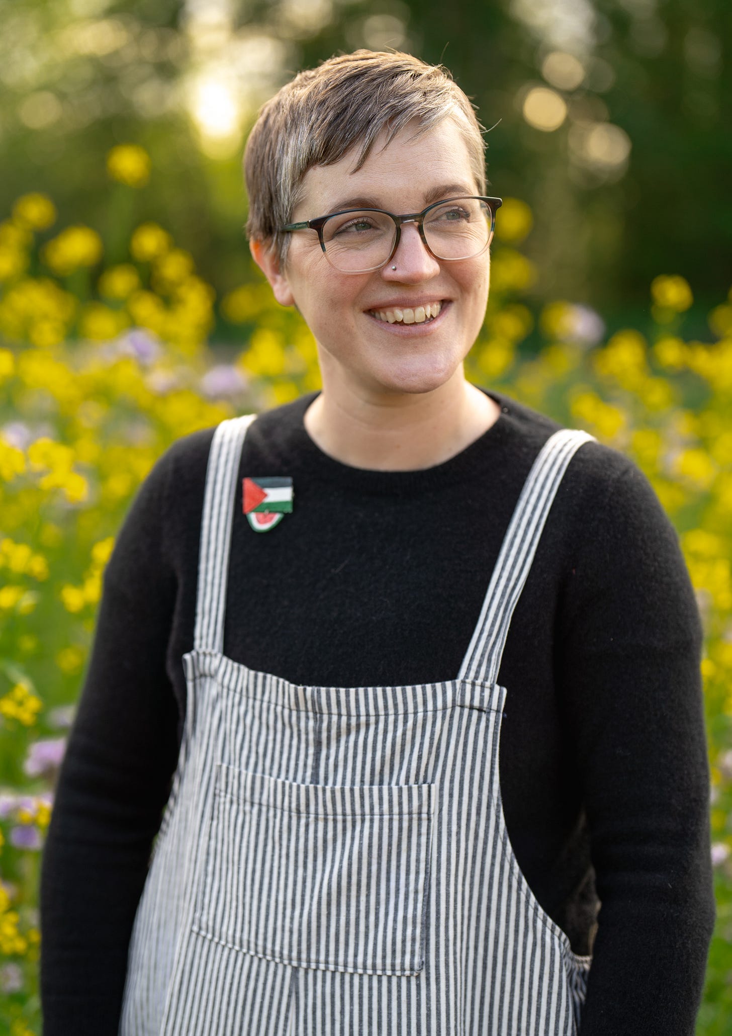 a white person with short graying hair and glasses is in front of a field of yellow flowers. they are smiling and wearing a black sweater with a small palestine pin a white person with short graying hair and glasses is in front of a field of yellow flowers. they are smiling and wearing a black sweater with a small palestine pin