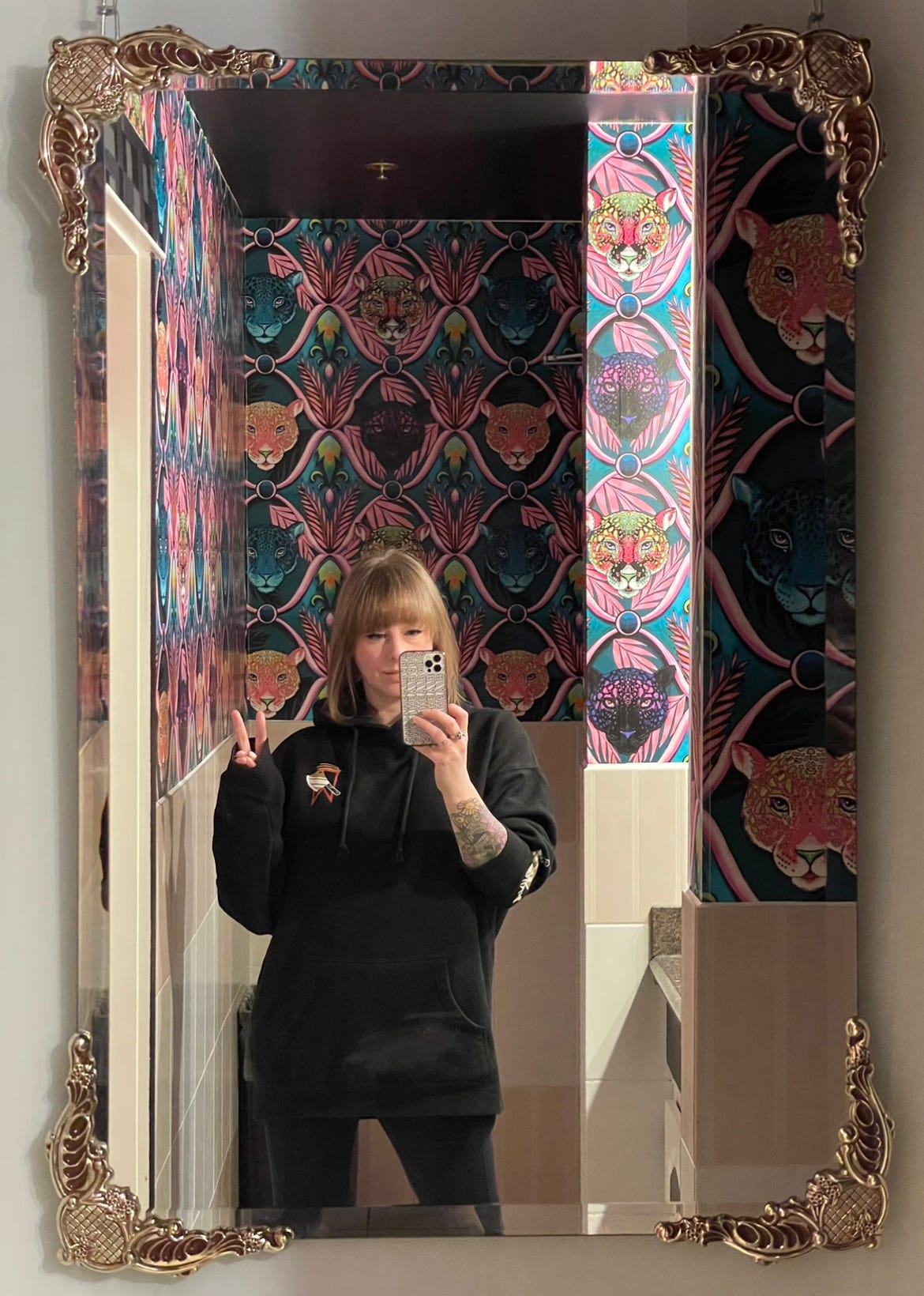 Image shows a blonde white woman wearing black clothes taking a picture of herself in an ornate mirror. Behind her is vibrant wallpaper with a design of jaguar faces.