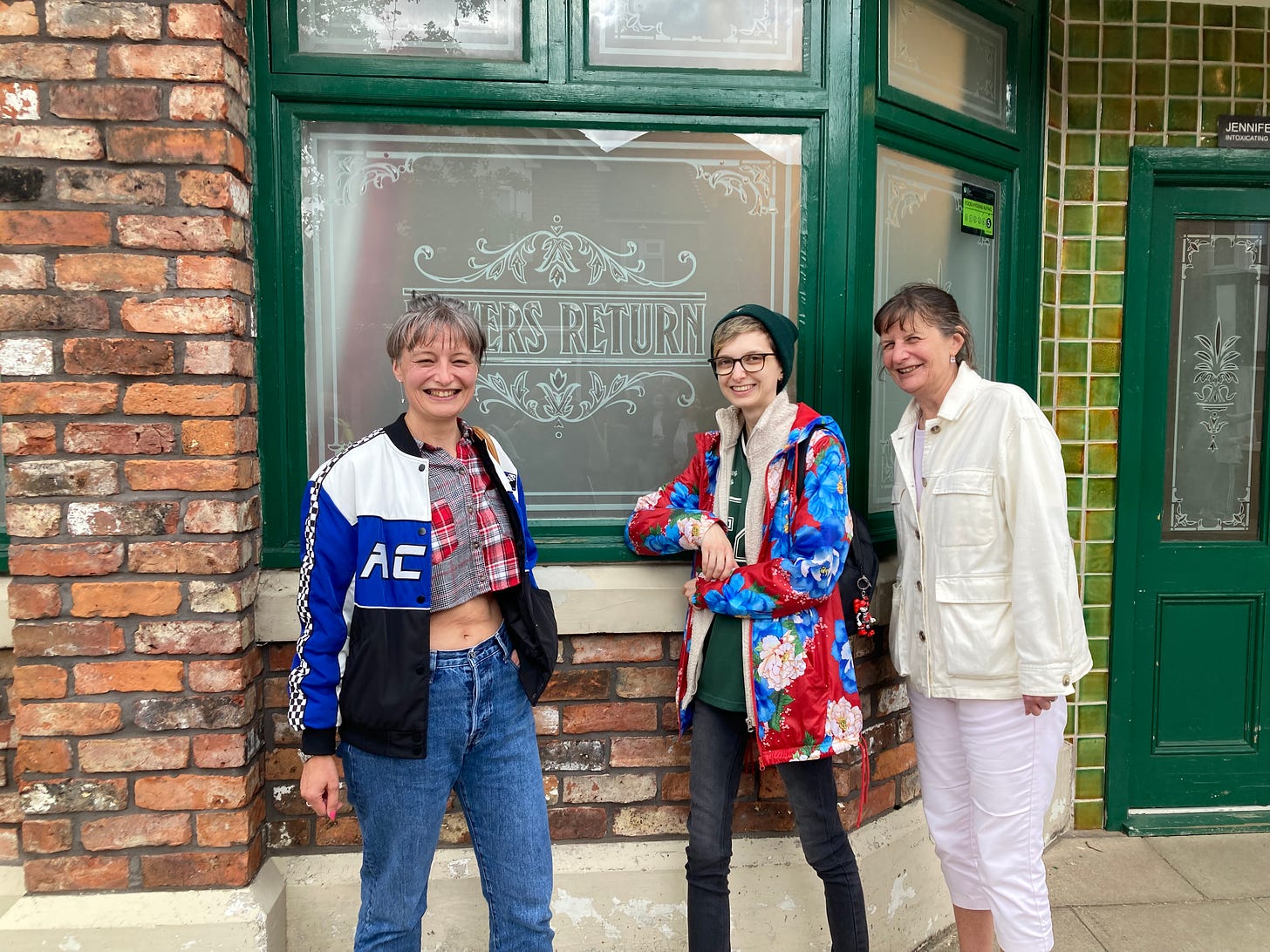 Photograph of Beck, Beck's mum and Beck's auntie all standing in front of the Rover's Return pub on the Coronation Street set. They are all smiling widely with teeth showing, in front of a window that is engraved with 'Rovers Returns'