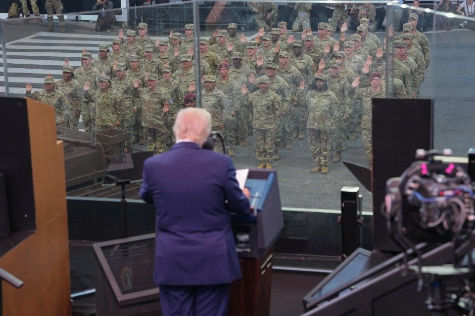 President Biden speaking at an Army 250th birthday celebration, viewed from behind, with soldiers taking an oath in the background.