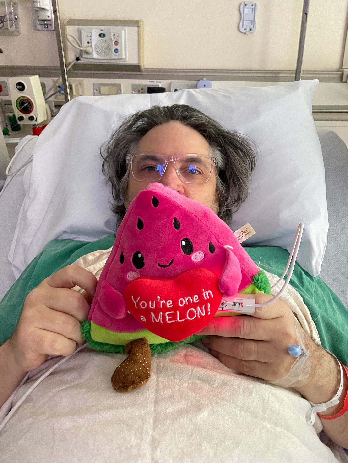 My dad lays in a hospital bed holding a plush watermelon toy that says You’re one in a melon.
