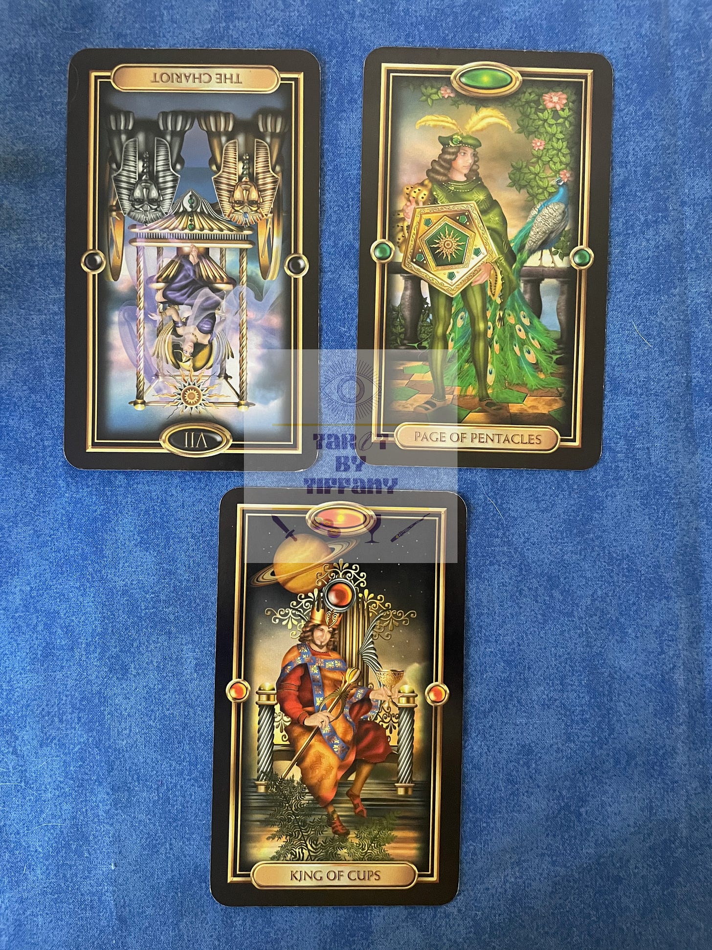 3-card reading with the Gilded Tarot. The cards are laid against a solid, cobalt blue cloth background. Top two cards (L-R): Chariot reversed, Page of Pentacles. Bottom card: King of Cups.