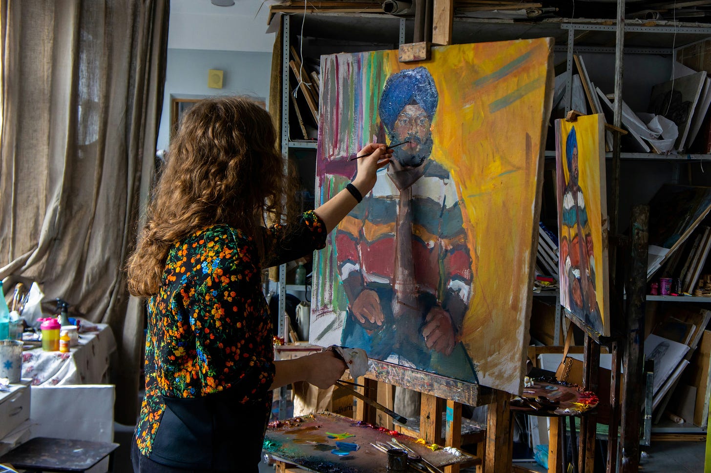 A woman with curly brunette hair paints a picture of a man wearing a blue turbin; white, green, yellow and red shirt, and green paints.