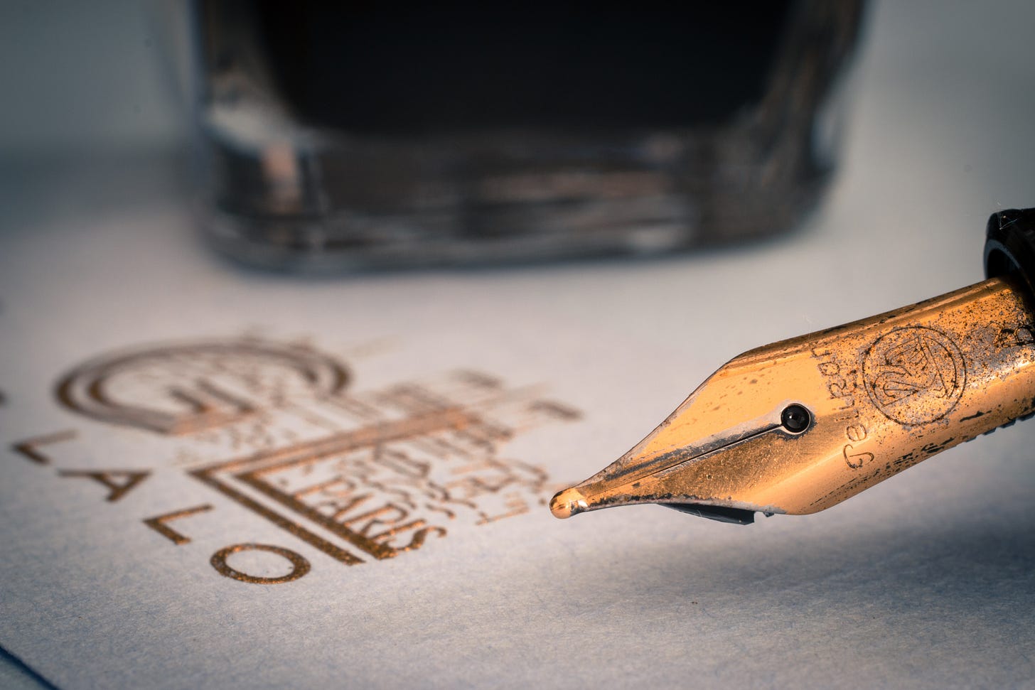 A close up image of a gold-nibbed fountain pen hovering over a fine fiberously textured paper stamped with the gold embossed logo of some unknown organization and an out of focus ink bottle in the background