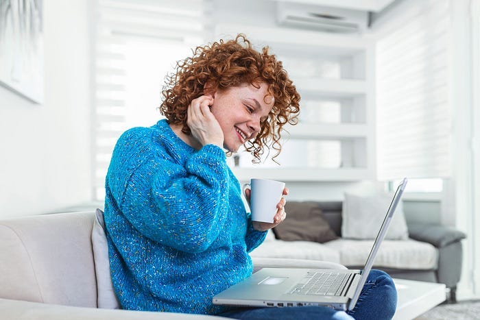 A happy woman in a blue sweater drinking coffee and writing on a laptop. A happy woman in a blue sweater drinking coffee and writing on a laptop.
