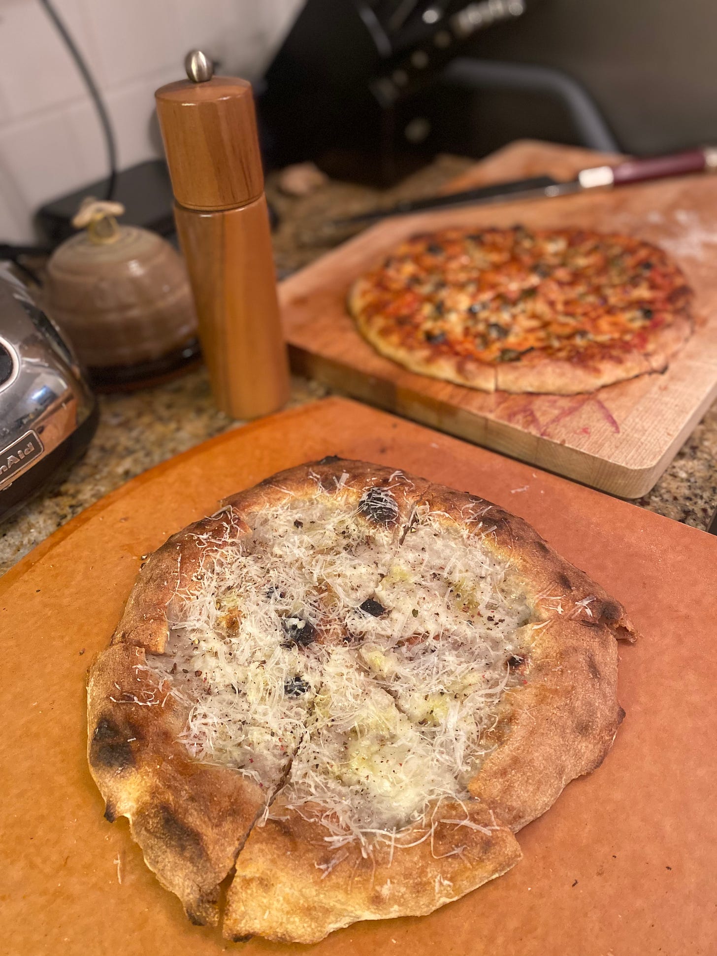 Two pizzas, one on the pizza paddle and the other on the cutting board in the background. In the background is a classic red sauce pizza with olives and mozza, and in the foreground is the cacio e pepe pizza described above. Next to it is a wood peppermill.