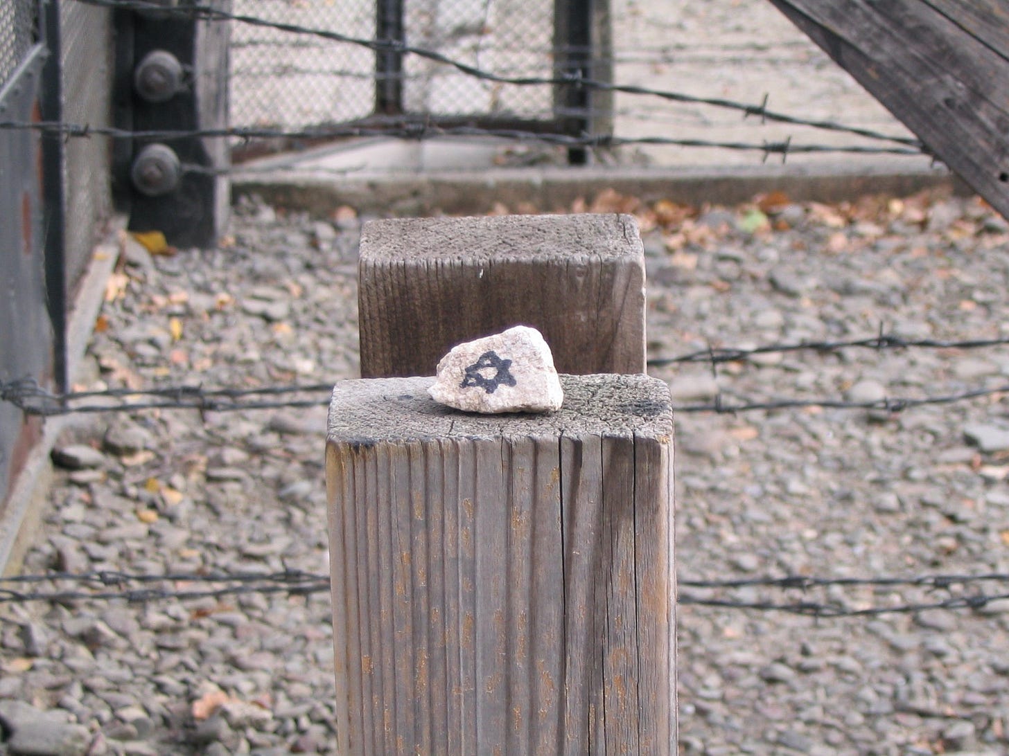 Photo of a small stone with a Star of David drawn on it on top of a fence post, barbed wire fence nearby, at the museum at Auschwitz.