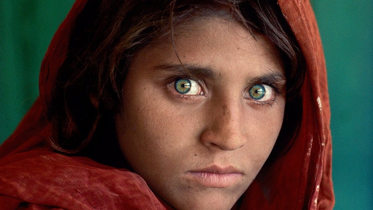 Close-up portrait of a young Afghan girl with striking green eyes and a red headscarf against a green background.