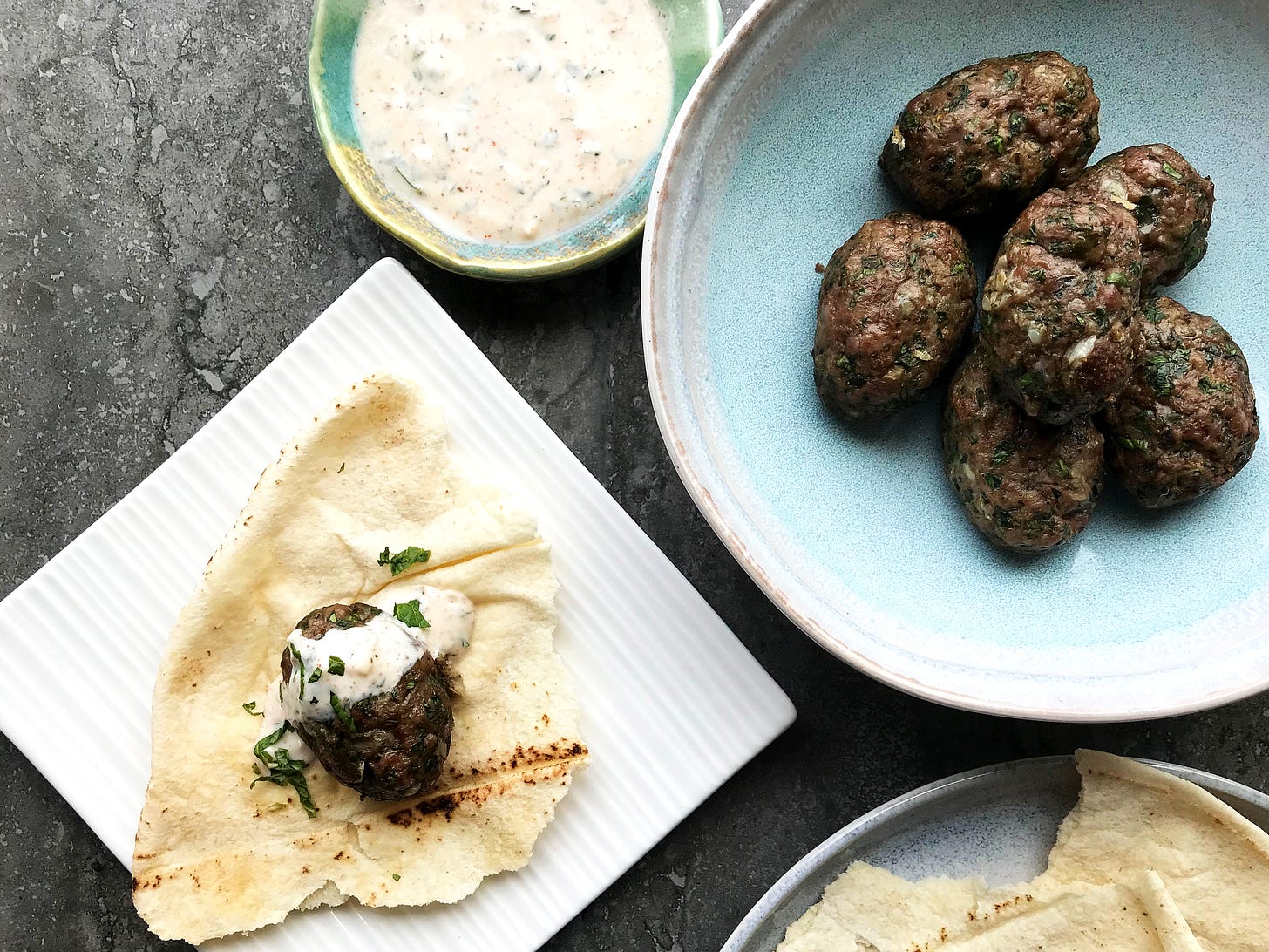 A bowl of kefta next to a small bowl of minted yoghurt sauce, a plate with a torn bit of pita on which sits a kefta, drizzled with the yoghurt sauce and sprinkled with chopped mint. Partially visible is a plate of torn pita.
