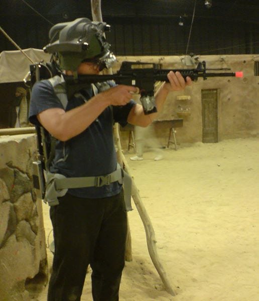 Me, wearing a helmet with mounted cameras, wearing a backpack with a laptop, holding a fake gun, while in a fake desert environment in a warehouse